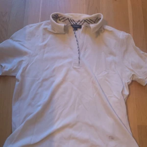 Vit Burberry pikétröja med dragkedja - Vit pikétröja från Burberry London i storlek L. Klassisk krage med Burberrys ikoniska rutiga mönster på insidan och längs dragkedjan. Kortärmad och tillverkad i bomull, perfekt för en clean och stilren look. Vet inte om den är äkta därav priset.