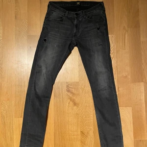 Mörk gråa Lee jeans - Säljer ett par svarta slim fit jeans från Lee, modell Luke. Jeansen har en smal passform, klassisk femficksdesign och diskreta slitningar framtill. Materialet är stretchigt. Har en liten fläck på höger ben men det går bort i tvätten.