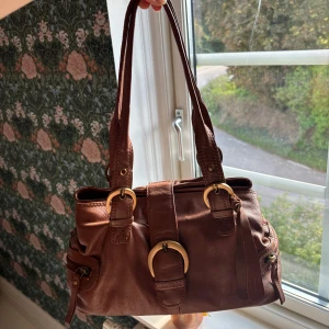 Brun handväska i äkta läder  - Snygg brun handväska  i äkta skinn med två axelband och guldfärgade spännen. Väskan har flera fack med dragkedja på sidorna och detaljerade sömmar. Perfekt storlek till fest då den är åt det mindre hållet. Skriv för mer frågor!!💕