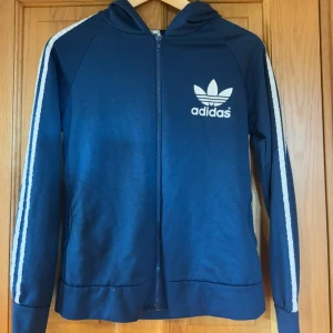 Blå hoodie med dragkedja - Säljer en blå hoodie med vit logga på bröstet och klassiska vita ränder längs ärmarna. Tröjan har huva, hel dragkedja och långa ärmar. Perfekt för en sportig och avslappnad stil. ‼️KOPIA‼️