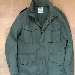 Grön  fieldjacket  från Dobber - Snygg militärgrön parkajacka från Dobber med fyra stora fickor framtill, hög krage och axelklaffar. Jackan har dragkedja och knappar framtill samt justerbar midja. Perfekt för dig som gillar en klassisk och stilren look.