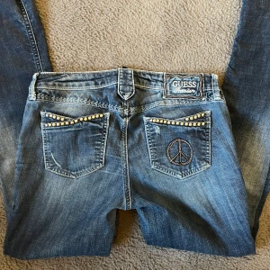 Guess Premium blå jeans - Snygga blå jeans från Guess Premium med boot cut. Byxorna har coola guldfärgade nitar på fickorna och ett broderat peace-märke bak. Insidan har blommigt mönster. Jeansen är avklippta längst ner. Jeansen är i storlek 28. De är i boot cut model