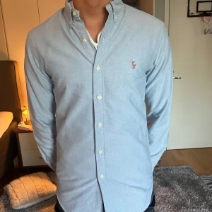 Ljusblå skjorta Polo Ralph Lauren - Snygg ljusblå skjorta från Polo Ralph Lauren i slim fit-modell. Skjortan har klassisk button-down krage, broderad logga på bröstet och vita knappar framtill. Tillverkad i mjuk bomull, perfekt för en clean och stilren look. Säljer denna snygga skjorta för den tyvärr har blivit för liten för mig. Nypris-1600kr/mitt pris-600kr!