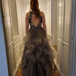 Mörkgrå balklänning med spetsdetaljer - Säljer en magisk mörkgrå långklänning med voluminös kjol i lager på lager av tyll. Toppen har transparent mesh med blommiga spetsapplikationer och en djup öppen rygg. Klänningen har ärmlös design och v-ringad front, perfekt för bal eller fest.