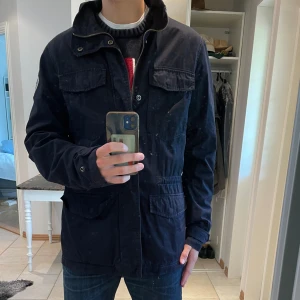 Marinblå fieldjacka från Napapijri - Säljer en mörkblå field jacka från Napapijri med fyra stora fickor framtill, dragkedja och tryckknappar. Jackan har hög krage och emblem på ärmen. Perfekt för dig som gillar stilren och funktionell design.