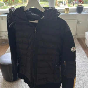 Svart cardigan - Svart cardigan från Moncler med stickade ärmar och quiltad front. Jackan har dragkedja framtill, fickor med tryckknappar och en extra ficka med dragkedja på ärmen. Klassisk Moncler-logga på ena ärmen. Perfekt för kyliga dagar.                  Pris kan diskuteras