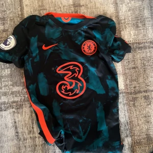 Chelsea FC Nike fotbollströja barn L / 147 158 - Chelsea FC matchtröja från Nike med svart och mörkgrönt abstrakt mönster, röda detaljer och klubbmärke på bröstet. Tröjan har korta ärmar, Dri-Fit-material och Premier League-märke på ena ärmen. Perfekt för dig som älskar fotboll och vill sticka ut.