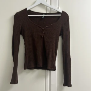 Brun ribbad långärmad topp Vero Moda  - Säljer en brun långärmad topp från Vero Moda med ribbad struktur och v-ringning. Toppen har dekorativa knappar framtill och är i en slim passform. Perfekt att styla med jeans eller kjol för en trendig look.
