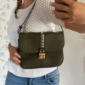 Olivgrön axelväska med gulddetaljer - Mörk olivgrön väska som har guldiga detaljer. Mycket bra skick utan låset som hoppar/ funkar inte ibland därav så billig. Ska gå att fixa. Har ett långt axelband som jag brukar göra kort som på bilden