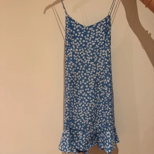 Blå blommig klänning från Zara - Supersöt blå klänning från Zara med vita småblommor, smala axelband som korsas i ryggen och volangkant nertill. Perfekt för varma dagar och ger en fräsch sommarkänsla. Lätt och luftig modell som sitter snyggt på kroppen. Är i storlek S. 