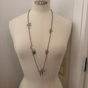 Långt silverfärgat halsband med fåglar - Unikt långt halsband i silverfärg med flera detaljerade fågelhängen och gnistrande stenar. Kedjan är tunn och smidig, och fåglarna ger halsbandet en cool vibe. 