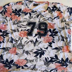 Blommig t-shirt med tryck från Bluewear - T-shirt från Bluewear i storlek XXL med blommigt mönster i rosa, svart och grått på vit botten. Stor svart siffra '79' tryckt framtill. Skön passform och korta ärmar, perfekt för dig som gillar sportig och färgglad stil.