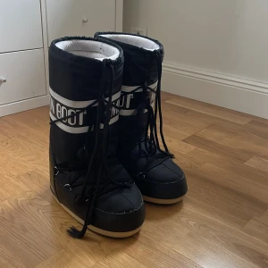 Svarta Moon Boot  - Säljer ett par klassiska svarta Moon Boot  med vit logga och snörning framtill. Skorna har en hög, vadderad design med platt sula och grovt mönstrad undersida för extra grepp. Perfekta för vintern och håller fötterna varma och torra.
