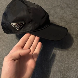 Svart keps från Prada - Snygg svart keps från Prada med ikonisk triangulär metallogo på sidan. Kepsen är tillverkad i glansig nylon och har en klassisk böjd skärm. Perfekt accessoar för en clean och lyxig streetstyle-look.