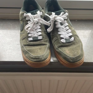 Nike Air Force 1 i grön mocka - Säljer ett par Nike Air Force 1 sneakers i grön mocka med vita snören och brun gummisula. Klassisk siluett med perforerad tå och låg profil. Swoosh-logga på sidan och AIR-detalj på hälen. Perfekta för dig som gillar streetwear och snygga detaljer.