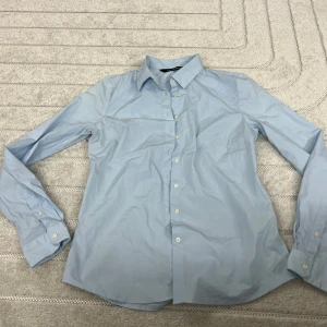 Ljusblå skjorta från Donna Karan - Klassisk ljusblå skjorta från Donna Karan med lång ärm och knappar framtill. Skjortan har en stilren krage och är tillverkad i bomull, perfekt för en clean och enkel look. Passar dig som gillar minimalistisk stil.