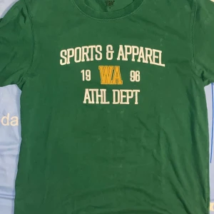 Grön t-shirt med tryck Sports & Apparel - Grön t-shirt med vit och gul text på bröstet: 'Sports & Apparel 1996 WA ATHL DEPT'. Klassisk rund hals och korta ärmar. Skön och avslappnad passform, perfekt för en sportig look. 