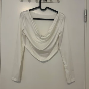 Vit långärmad omlottopp med drapering - Snygg vit långärmad topp med draperad omlottringning och spetsig nederkant. Toppen har en stilren look och är tillverkad i ett mjukt, stretchigt material som sitter skönt på kroppen. Perfekt för dig som gillar trendiga och enkla plagg med en twist.