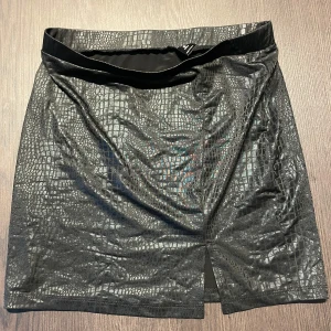 Svart kjol med krokodilmönster - Snygg svart kortkjol med glansig krokodilstruktur och liten slits framtill. Kjolen har en tight passform och är perfekt för dig som vill sticka ut med en cool look. Materialet är syntet och ytan har en läcker glans.