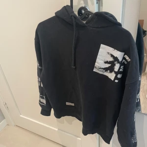 Svart hoodie från Stay Out - Svart hoodie från Stay Out med tryck i vitt och grått på bröstet och ärmarna. Tröjan har huva med snörning och en avslappnad passform. Perfekt för dig som gillar streetwear och vill sticka ut lite extra.