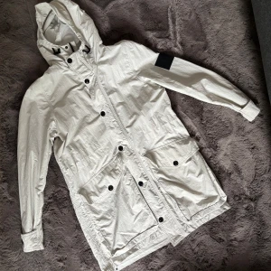 Vit parkajacka från Peak Performance - Snygg vit parkajacka från Peak Performance med huva, stora fickor framtill och svarta knappar. Jackan har en svart patch på ärmen och är tillverkad i ett lätt och slitstarkt material. Perfekt för dig som vill ha en clean och stilren look.