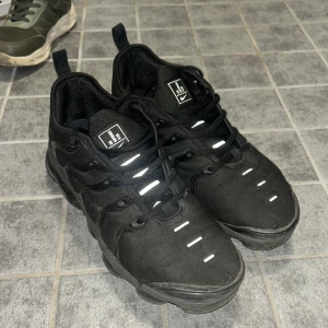 Nike Air Vapormax Plus Svarta Sneakers - Svarta Nike Air Vapormax Plus sneakers med reflekterande detaljer och ikonisk bubblig sula. Ovandel i textil och syntet, diskret Swoosh på sidan och Vapormax Air-märke bak. Grymt snygga och bekväma skor med snörning och modern design.