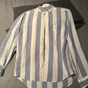 Randig linneskjorta från GANT - Snygg skjorta från GANT i linne med breda vertikala ränder i blått och vitt. Klassisk krage, bröstficka med broderad logga och knäppning framtill. Perfekt för en chill och stilren look.