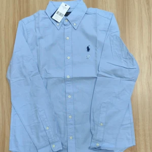 Ljusblå skjorta från Polo Ralph Lauren - Helt oanvänd! Ny Klassisk ljusblå skjorta från Polo Ralph Lauren med button-down krage och broderad logga på bröstet. Skjortan har lång ärm, vita knappar och är tillverkad i mjuk bomull. Perfekt för en clean och stilren look.🏖️🧊🌊