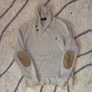 Beige kofta från Ralph lauren  - Snygg grå tröja från Polo Ralph Lauren med klassisk krage och tre knappar vid halsen. Tröjan har långa ärmar med mockadetaljer på armbågarna och den ikoniska broderade loggan på bröstet. Perfekt för dig som gillar stilren och tidlös design.