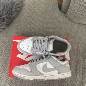 Nike Dunk Low vit/grå sneakers - Säljer ett par Nike Dunk Low sneakers i vitt och ljusgrått. Skorna har klassisk siluett med rund tå, snörning och platt sula. Ovandelen är i skinn och textil, med ikonisk Swoosh på sidorna. Perfekta för dig som gillar stilrena och trendiga sneakers.