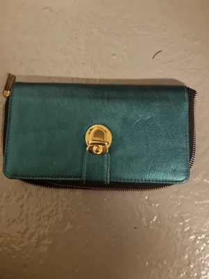 Turkos plånbok med gulddetalj - Snygg turkos plånbok i skinn med guldfärgat spänne framtill och dragkedja runtom. Insidan har ett coolt geometriskt mönster i svart och vitt, samt flera fack för kort och mynt. Perfekt för dig som vill ha något både praktiskt och stilrent.