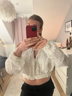 Vit broderad blus med knyt - Superfin vit blus i bomull med broderade hålmönster och offshoulder-modell. Toppen är croppad med resår i midjan och har långa puffiga ärmar samt knytning framtill. Perfekt för vår och sommar när du vill ha en luftig och söt look.