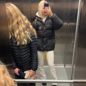 Svart dunjacka från Fjällräven - Säljer en svart dunjacka från Fjällräven med gula snören och klassisk logga på bröstet. Jackan har huva, stora fickor med knappar och dragkedja framtill. Perfekt för kalla dagar och riktigt skön passform med lite oversized känsla.