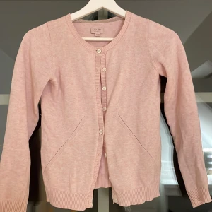 Ljusrosa kofta från Noa Noa Miniature - Säljer en söt ljusrosa kofta från Noa Noa Miniature i storlek 140/10Y. Koftan har rund hals, långa ärmar och knappar framtill. Ribbstickade muddar vid ärmslut och nederkant ger en snygg finish. Perfekt att slänga över en topp när det är lite kyligt.