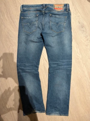 Blå Diesel jeans med slitningar - Säljer ett par vintage blå jeans från Diesel med snygga slitningar framtill och tydliga bakfickor med V-formad söm. Jeansen har normal passform, knappgylf och är tillverkade i robust denim. Perfekt för dig som gillar en avslappnad och cool stil.