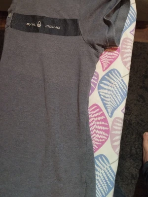 Grå piké från Sail Racing - Säljer en stilren grå pikétröja från Sail Racing med svart detalj och logga på bröstet. Tröjan har korta ärmar, klassisk krage och knappar vid halsen. Materialet känns som bomull och ger en avslappnad men ändå sportig vibe.