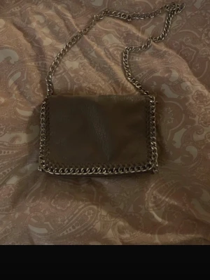 Grå axelväska med kedja från Tiamo - Säljer en grå axelväska från Tiamo med silverfärgad kedja som axelrem och runt kanten. Väskan är gjord i skinnimitation med struktur och har dragkedja både invändigt och på baksidan. Perfekt storlek för det viktigaste. Den har lite skavanker, se bild 4