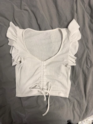 Vit croppad topp med volangärmar - Säljer en vit croppad topp med volangdetaljer på ärmarna och snygg snörning framtill. Toppen har v-ringning och är tillverkad i ett ribbat material som ger en skön och stretchig känsla. Perfekt till jeans eller kjol för en trendig look.