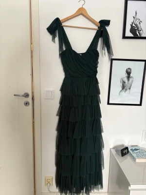Mörkgrön långklänning med volanger - Säljer en mörkgrön långklänning med flera lager volanger och knytband på axlarna. Klänningen är gjord i tunn mesh och har omlott-detaljer upptill. Perfekt för dig som vill ha en romantisk och flowy look. Storlek 10 storbritannien och 38 sverige.