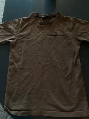 Brun t-shirt från Sail Racing - Brun t-shirt från Sail Racing med diskret logga på bröstet och ett tryck på ryggen. Klassisk rund hals och korta ärmar. Materialet är mjuk bomull som känns skönt mot huden. Perfekt för dig som gillar enkel och stilren design jag har även en sail racing påse till själva tshirten. .