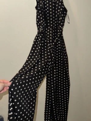 Svart prickig byxdress med vida ben - Säljer en svart byxdress med vita prickar och vida ben. Byxdressen har ärmlös design, knappar vid axeln och ett lätt, flowy tyg som känns riktigt skönt. Perfekt för dig som gillar en lekfull men ändå stilren look.