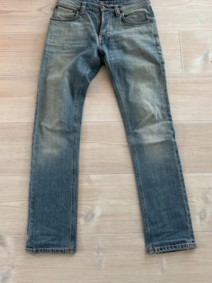 Blå jeans från Nudie Jeans, stl 28/30 - Säljer ett par klassiska blå jeans från Nudie Jeans med snygga slitningar och kontrastsömmar. Modellen har rak passform och fem fickor, med Nudie Jeans logga på bakfickan. Materialet är robust denim i bomull och färgen är ljusblå med tvättad look. Sitter som w27