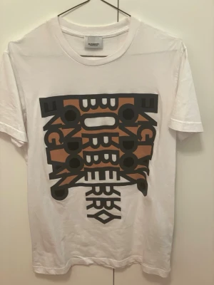  Burberry t-shirt - Säljer en vit t-shirt från Burberry med ett stort grafiskt tryck i svart och brunt på bröstet. Trycket har texten 'Burberry London England' i en cool, modern design. T-shirten har rund hals och korta ärmar, perfekt för dig som gillar streetstyle och designerplagg.
