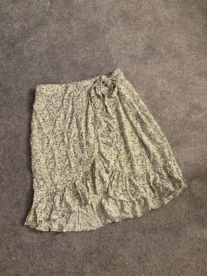 Blommig omlottkjol från Vila - Säljer en söt omlottkjol från Vila i storlek 34. Kjolen är ljusbeige med små blå blommor och har volangkant nedtill. Den knyts i midjan och är perfekt för vår och sommar. Materialet är mjukt och lätt och är endast använd några få gånger. 