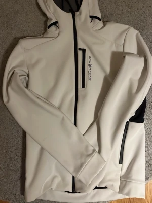 Vit Sail Racing vindjacka Gore-Tex L - Snygg vit vindjacka från Sail Racing med svarta detaljer och huva. Jackan har flera dragkedjor, bröstficka och är tillverkad i Gore-Tex Infinium-material som är både vindtätt och vattenavvisande. Perfekt för dig som gillar stil och funktion.