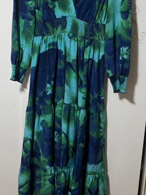 Blommig långklänning i blått och grönt - Säljer en maxiklänning med blommigt mönster i blå och gröna toner. Klänningen har lång ärm med smock vid ärmslut och midja, samt v-ringad hals. Materialet känns lätt och flowy.