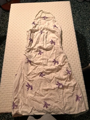 Vit långklänning med lila broderi - Säljer en vit långklänning med lila broderade blommor. Klänningen har halterneck och dragkedja i ryggen. Materialet känns som bomull och modellen är ärmlös med en lätt A-linjeform. Perfekt för sommarens varma dagar.