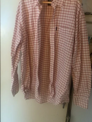 Vit rutig skjorta från Sebago - Vit skjorta från Sebago med röda rutor, klassisk krage och bröstficka. Skjortan har långa ärmar och knäppning framtill. Perfekt för dig som gillar en clean och stilren look med lite preppy känsla.