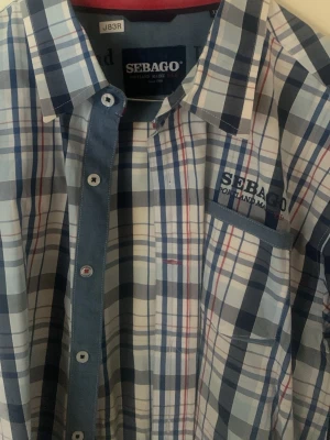 Rutig långärmad skjorta från Sebago - Säljer en snygg rutig skjorta från Sebago i blå, vit och röda toner. Skjortan har klassisk krage, knappar framtill och en bröstficka med broderad logga. Perfekt för dig som gillar en chill och stilren look.