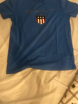 Blå GANT t-shirt med tryck - Snygg blå t-shirt från GANT med stort tryck framtill av logga och sköld med stjärnor och ränder. Klassisk rund hals och korta ärmar. Tillverkad i mjuk bomull som känns skön mot huden. Perfekt för dig som gillar stilrena och sportiga plagg.